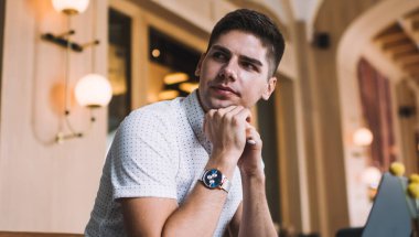 Düşünceli, genç bir erkek. Ellerine yaslanıp çenesine dokunarak yaratıcı bir iş yerinde çalışırken bilgisayarını kullanıyor.