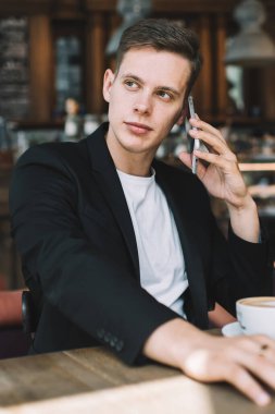 Genç, iyi giyimli, cep telefonuyla konuşan ve şık bir kafede bir fincan kahveyle otururken yüzünü çeviren bir erkek.