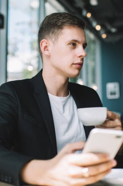 Siyah ceketli, beyaz tişörtlü, kahve içen ve kafeteryada tam pencereli bir şekilde dikkatle bakan konsantre genç adam.