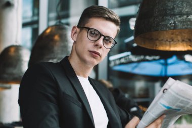 Gözlüklü, şık giyinmiş bir adam, elinde gazete ve bir fincan sıcak içecekle şık bir kafede otururken kameraya bakıyor.