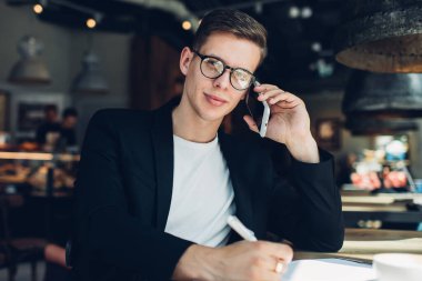 Kendine güveni tam bir adam, modern bir restoranın masasında oturmuş, iş projesinde not defterine planlar yazıyor ve cep telefonuyla konuşuyor.