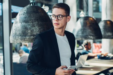 Gözlüklü, siyah şık takım elbiseli, cep telefonu kullanan ve şık bir kafede masaya yaslanırken yüzünü çeviren genç bir erkek.