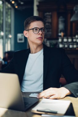 Siyah ceketli ve gözlüklü, kafeteryada kahve içip gözlerini kaçıran ciddi bir erkek işçi.