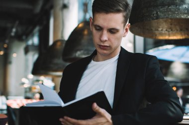 İyi giyimli siyah takım elbiseli bir erkek, teneffüste masanın yanında oturup kitap okuyor ve modern kafeteryada dinleniyor.