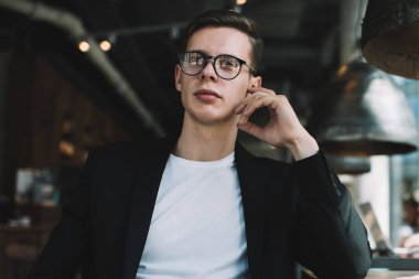 Ciddi, gözlüklü, dizüstü bilgisayarlı genç bir adam ara sırasında modern kafede bir fincan kahve içerken kameraya bakıyor ve düşünüyor.