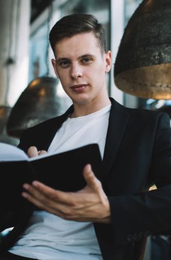 İyi giyimli, kendine güvenen bir erkek girişimci kafede oturuyor ve proje üzerinde çalışırken notlar alıyor ve kameraya bakıyor.