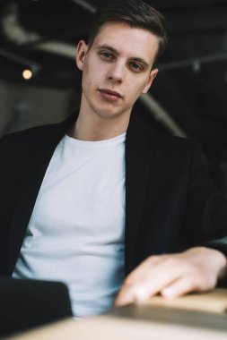 Kendine güvenen genç işadamı ellerini masaya koyup kameraya bakarken modern kafeteryada bir fincan kahveyle dinleniyor.