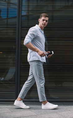 Siyah şeffaf bölmenin yanından geçerken cep telefonuyla dolgu malzemesi içinde yürüyen ve kameraya bakan genç bir erkek.