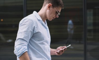 Gündelik kıyafetli ve güneş gözlüklü bir erkeğin cep telefonuyla internette gezinirken kaldırımda el ele tutuşup güneş ışığının altında duruşu.