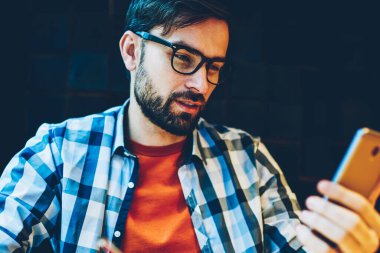 Dalgın sakallı öğrenci Not defteri smartphone aygıtından metin bilgileri yeniden yazma. Web sitesinde telefon haber okuma ve kafede oturan Not defterinizdeki notlar yapma gözlük hipster adam