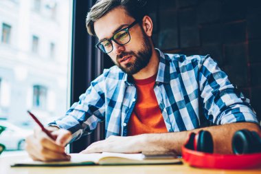 Dalgın sakallı öğrenci ödev Not defteri alanı coworking eğitim aşağı yazma. Akıllı hipster adam ders notları not defterinde kafede sınava hazırlık sırasında yapma gözlük