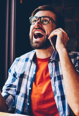 Ağlama ve telefon görüşmesinde smartphone aygıtında savunarak rahat giyimli kızgın sakallı genç adam. Konuşacak coworking içinde oturan modern cep telefonuna bağıran gözlük rahatsız hipster adam