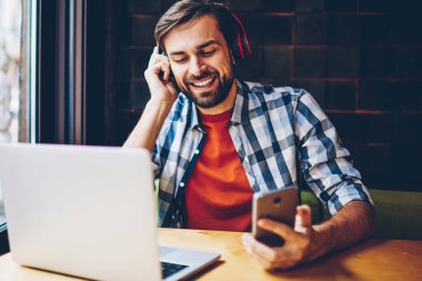 Mutlu sakallı genç modern kulaklık smartphone aygıtında indirilen çalma listesinden müzik dinleme adam. Dijital dizüstü bilgisayar başında oturan radyo web sitesinde ses şarkı zevk neşeli erkek blogger
