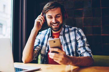 Modern kulaklık dinlemek için neşeli sakallı hippi adam şarkı müzik uygulaması seçme smartphone için bluetooth ile bağlantılı. Olumlu erkek blogger cep çalar ses tadını çıkarırken sohbet