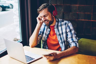Smartphone için Bluetooth kaygısız sakallı genç adam şarkı şarkı dinleme ses modern kulaklık girişi sırasında bağlı. Neşeli erkek blogger dijital dizüstü bilgisayar kafede oturan müzik keyfi