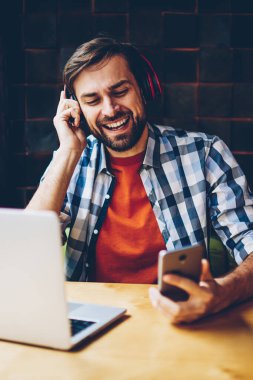 Pozitif sakallı genç adam favori ses çalma listesinden şarkıyı modern Kulaklığımda Smartphone'da indirilebilir. Mutlu hipster adam dinleme müzik radyo oyuncu laptop oturan telefonu