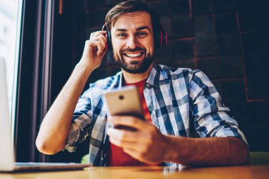 Mutlu sakallı erkek blogger smartphone için Bluetooth kulaklık uygulamada müzik dinleme şarkıları bağlı iken kameraya gülümseyen portresi. Neşeli hipster adam zevk çalma listesi