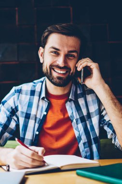 Smartphone üzerinde arama ve notlar not defteri alanı coworking oturan yapım sırasında kameraya gülümseyen başarılı sakallı öğrenci portresi. Pozitif hipster adam telefon aygıtı üzerinde iletişim