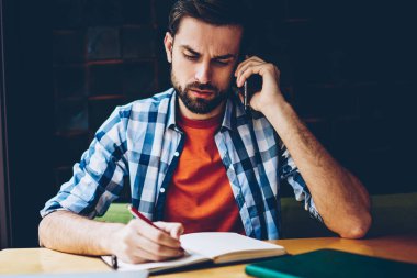 Konsantre hipster adam coworking masada oturan smartphone üzerinde konuşma sırasında duydum Not Defteri metin bilgileri yazma rahat forması giymiş. Cep üzerinde calling düşünceli öğrenci