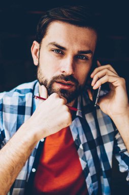 Ciddi sakallı genç adam portresi smartphone aygıtında çağrılırken kameraya bakarak rahat şık gömlek giymiş. Yakışıklı erkek blogger işleci ile telefon görüşmesi sırasında iletişim