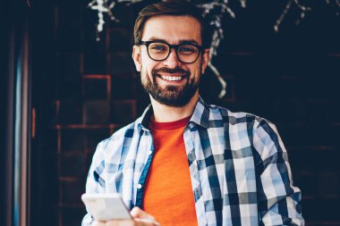 Mutlu portre alan coworking içinde smartphone aygıt ayakta kısa mesaj yazarken kameraya gülümseyen genç adam sakallı. Pozitif hipster adam Online sosyal ağlar üzerinde telefon sohbet