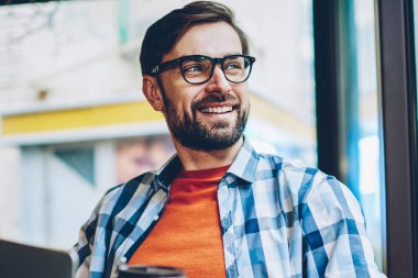 Gülümseyen hayal pencereden dışarı bakıyor ve öğle arasında Web sitesi geliştirmek için yaratıcı fikirler üzerinde düşünme genç adam sakallı. Düşünceli neşeli erkek blogger ile netbook aygıt çalışma gitmek için kahve