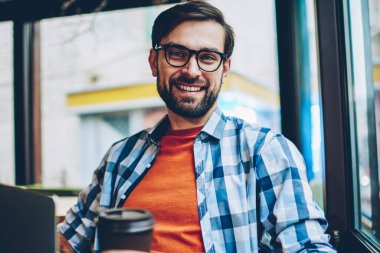 Yarım uzunluk portre gitmek başarılı sakallı genç adamın gözlük kamera ile kahve oturan gülümseyen vizyon düzeltme için coworking içinde eller. Pozitif hipster adam rahat gömlek giymiş