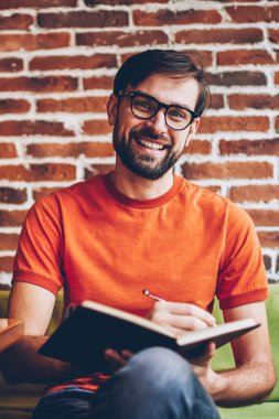 Alan coworking içinde modern netbook oturan işlem planı başarılı sakallı genç adam Not Defteri metin bilgileri yazarken kameraya gülümseyen Eyewear yarım uzunluk portresi