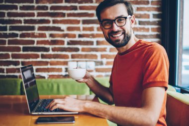 Portre kahve elinde tutan ve modern dizüstü bilgisayar yazılımı güncelleştirmek için 4 g bağlıyken kameraya gülümseyen başarılı sakallı genç adam Freelancer internet kafede oturan