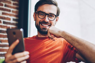 Başarılı sakallı erkek blogger smartphone aygıt ücretsiz 4 g internet bağlantısı üzerinden app karşıdan yüklerken kameraya gülme portresi. Pozitif hipster adam cep üzerinde online ödeme yapma