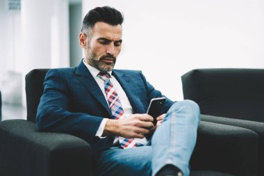 Dalgın olgun girişimci giyinmiş formal giyim finansal haber yolu ile 4 g Smartphone cep telefonu ile internet sitesinde okumak içinde ofis iç koltukta oturan internet. Gurur CEO'su posta kontrol konsantre