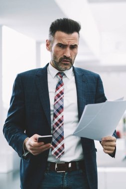 Deneyimli icra müdürü yarım uzunluk portre ofiste ayakta muhasebe raporları denetlerken kameraya bakarak formal giyim giymiş. Elinde telefon ile kendine güvenen gurur ceo