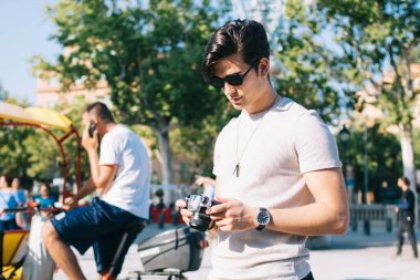 Ciddi, beyaz profesyonel fotoğrafçı güneşli bir günde şehir meydanında fotoğraf çekmek için kamerayla yürüyor. Güneş gözlüklü hippi turist tatil beldelerini geziyor.