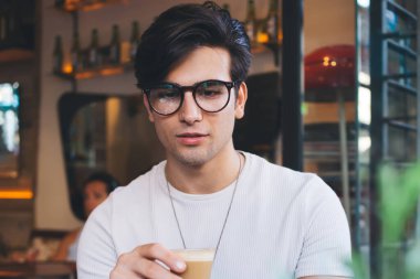 Ciddi yakışıklı adam dijital göçebe kahve içiyor ve dizüstü bilgisayarda video seyrediyor. Kafenin içinde uzaktan çalışıyor. Düşünceli hippi adam internet kursuna Webinar bardakla çalışıyor.