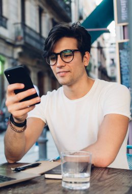 Gülümseyen yakışıklı erkek nüfuzlu kişi, akıllı telefon 4G internet bağlantısıyla sosyal ağlarda içeriği paylaşıyor. Kafe terasında dizüstü bilgisayar ve cep telefonu kullanarak çalışan gülümseyen hippi adam serbest çalışıyor.