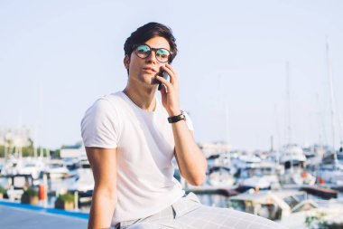 Moda giysileri ve gözlüklü yakışıklı gezgin uluslararası bağlantıları kullanarak akıllı telefon görüşmesi yapıyor genç hippi, deniz altyapısında akıllı telefondan konuşuyor.