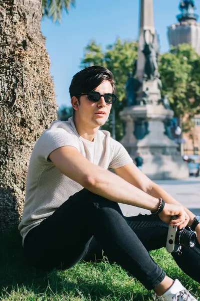 Parkta oturup hobisine zaman harcayan neşeli, yakışıklı hippi portresi. Elinde güneş gözlüklü gezgin adam. Tatilde şehri ziyaret ederken fotoğraf çektiği için elinde kamera var.