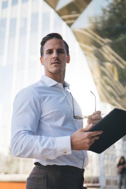 Resmi giysileri ve tablet ve gözlükleriyle ciddi bir girişimci modern şehir bölgesinde dikilirken dikkatlice kameraya bakıyor.