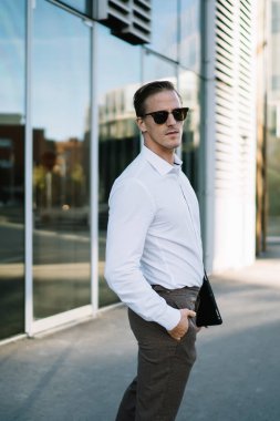 Şık kıyafetler ve güneş gözlüğü takan kendine güvenen erkek girişimci güneşli bir günde cam bina duvarının arka planında tabletle el ele duruyor ve kameraya bakıyor.