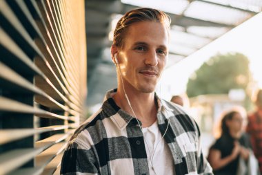 Bin yıllık olumlu beyaz erkek şehir merkezinde duruyor cep telefonu uygulaması ve aksesuar ile müzik dinliyor, gülümseyen adam güneşli bir günde 4G bağlantısı ve akıllı telefondan çevrimiçi radyodan keyif alıyor.