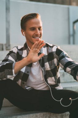 Gülümseyen beyaz erkek blogcu video görüşmesinde el sallıyor. Modern akıllı telefon açık havada oturuyor. Kulaklıklı mutlu erkek gezgin cep telefonuyla selfie için poz veriyor.