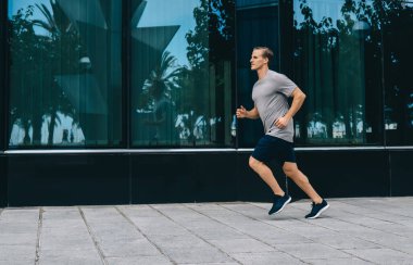 Kentteki kardiyo eğitimi sırasında koşan kararlı erkek koşucunun yandan görünüşü, fiziksel yoğunluk üzerinde yoğunlaşmak için ısınma sırasında dayanıklılık egzersizi yapan beyaz bir adam.