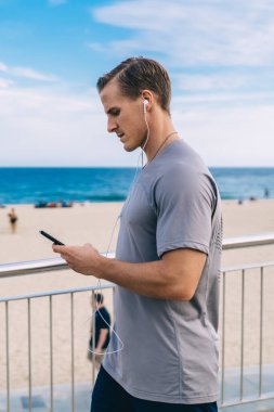 Elektronik kulaklıklı sporcu çevrimiçi mesajlaşma için modern cep telefonu aygıtını kullanıyor, kulaklık takıp kulaklık takan Kafkas formundaki antrenör medya mobil uygulamasından müzik seçiyor.