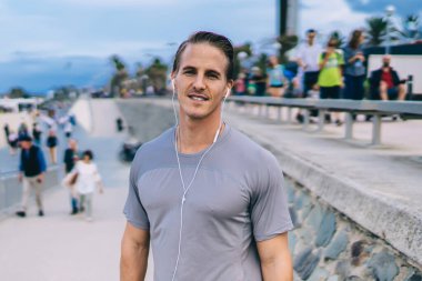 Kulaklıklı yakışıklı erkek koşucunun yarım boy portresi antrenman sırasında dinleniyor cep telefonundan radyo ve müzik listesi şarkıları dinliyor genç sporcu antrenmanda kameraya bakıyor.