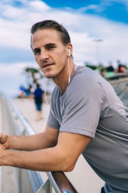 Gülümseyen 20 'li yaşlardaki yakışıklı hipster adamın spor molasında dinlenen portresi. Uygulamadan müzik dinlemek için cep telefonu kullanıyor. Pozitif sportmen açık havada akıllı telefondan koşmak hakkında podcast yapmaktan hoşlanıyor.