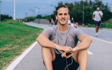Yakışıklı koşucunun portresi asfalt yolda koşmak ve kameraya bakmak için dinleniyor, elektronik kulaklıklı kaslı sporcu ses eğitimi almış müzik podcast 'i dinliyor ve poz veriyor.