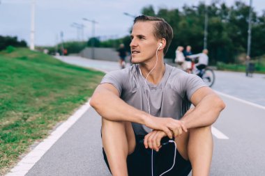 Elektronik kulaklıklı beyaz bir adam asfalt yolda dinleniyor. Kardiyo eğitimi için ses tavsiyesi dinliyor. 30 'lu yaşlarda kulaklıklı bir erkek antrenman sırasında medya uygulaması kullanıyor.