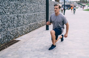 Beyaz tenli atletik kulaklıklı, elektronik kulaklıklı sokak antrenmanı sırasında çömelmiş, 30 yaşında kulaklıklı bir sporcu bacak kaslarını esnetirken müzik yayını dinliyor.
