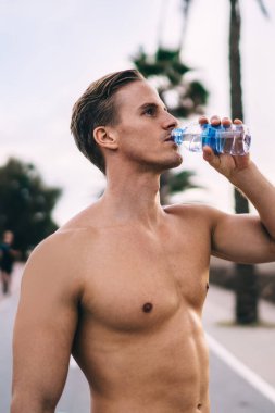 Sokakta antreman yaptıktan sonra mükemmel vücut suyu içen yakışıklı beyaz erkek koşucu, üstsüz sporcu, kardiyo çalıştıktan sonra susadıktan sonra dinlenmek için mola veriyor.