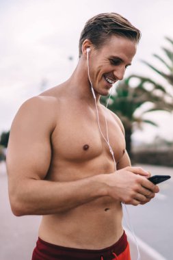 Seksi vücutlu mutlu beyaz atlet müzik podcast 'i dinlemek için akıllı telefon medya uygulaması kullanıyor, neşeli üstsüz vücut geliştirici mesajları yazıyor ve blog takipçileri sportif cevaplar yazıyor.